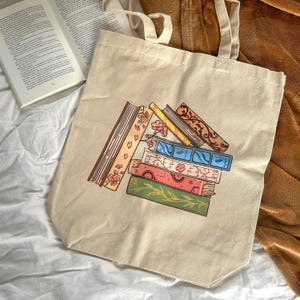Op de afbeelding: Beige canvas draagtas met een kleurrijke illustratie van een stapel boeken. De boeken hebben bloemen-, geometrische en bladpatronen in roze, blauw, oranje en groen. De tas heeft crèmekleurige handvatten en ligt naast een open boek.