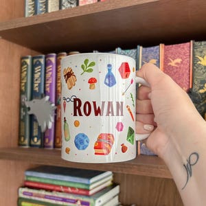 Op de afbeelding: Witte keramische mok met de naam "Rowan" in het rood, versierd met kleurrijke fantasie-thema illustraties. De mok wordt vastgehouden voor een boekenplank vol boeken.