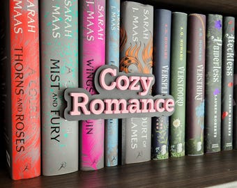 May include: A bookshelf displays a collection of books with colorful spines. The books are arranged in a row, with titles and author names visible. A pink and gray sign that says "Cozy Romance" is in front of the books.