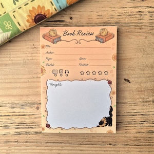 May include: A rectangular peach-colored notepad titled Book Review with illustrations of books, yarn balls, and sunflowers. It features lines for title, author, pages, genre, started, and finished dates, plus icons for reading format and five stars for rating.