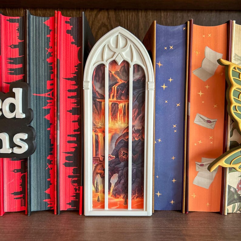 May include: A white, arched, gothic-style window frame insert featuring a printed scene of a volcanic landscape with flowing lava and two small dragons. The insert is positioned between several hardcover books on a shelf.