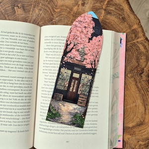 Op de afbeelding: Decoratieve boekenlegger met een gedetailleerde illustratie van een boekwinkel onder een kersenbloesemboom. De boekenlegger heeft een zwarte rand en ligt op de pagina's van een boek.