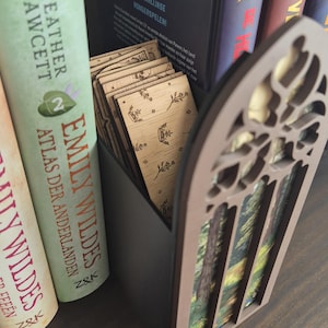 May include: A dark grey rectangular container holding several light-coloured wooden rectangular cards, each featuring a small engraved floral design. The container has a brown, arched, laser-cut decorative front panel with a forest scene print.