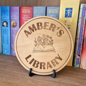 Op de afbeelding: Een rond houten bord met de tekst "Amber's Library" in een decoratief lettertype. Het bord is gemaakt van bruin hout en heeft gegraveerde bloemen en een open boek.