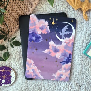 Può includere: Una carta rettangolare con un'illustrazione fantasy. Presenta una silhouette di drago su una luna crescente, circondata da nuvole rosa e viola e stelle. È visibile anche una statuina di drago dorato.