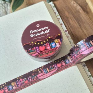 Op de afbeelding: Een rol washi tape met een bordeauxrode achtergrond en de tekst "Romance Bookshelf". De tape heeft een boekenplankontwerp met harten, boeken en feeërieke lichtjes. De tape is 15 mm x 10 m.