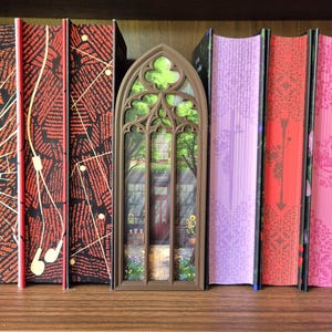 May include: A brown, arched, gothic-style book nook insert featuring a miniature bookstore scene with a storefront, a door, and greenery, positioned between two black books with red patterns and two books with pink and purple covers.