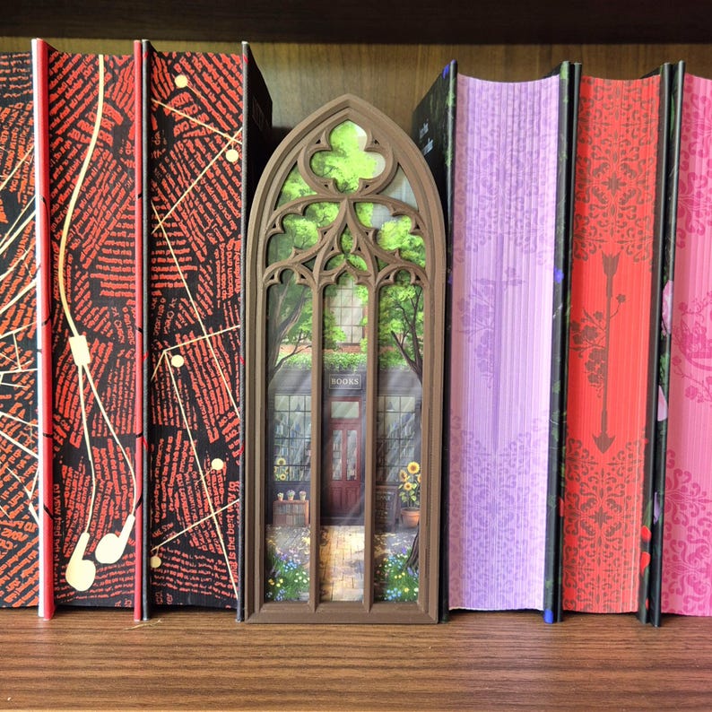 May include: A brown, arched, gothic-style book nook insert featuring a miniature bookstore scene with a storefront, a door, and greenery, positioned between two black books with red patterns and two books with pink and purple covers.