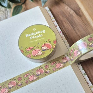 Op de afbeelding: Een rol washi tape met een "Hedgehog Picnic" ontwerp. De tape heeft een herhalend patroon van roze egels, boeken en picknickmanden op een groene achtergrond. De rol is 15 mm x 10 m.