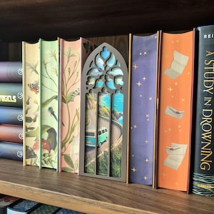 May include: A dark brown, arched, gothic-style wooden book nook insert featuring a miniature coastal scene with a turquoise van driving along a winding road by the ocean, positioned between several books with illustrated spines on a wooden shelf.