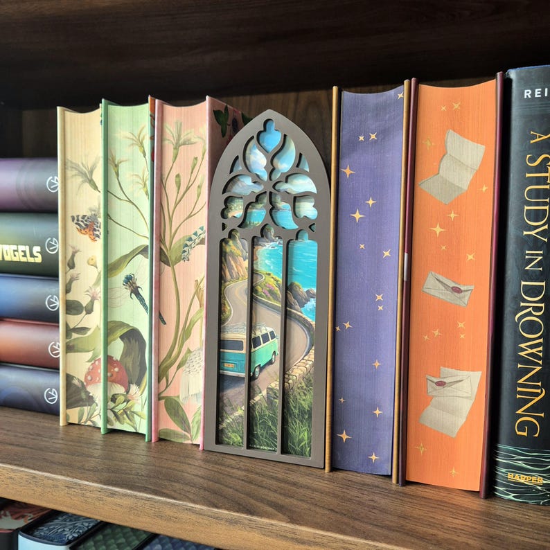 May include: A dark brown, arched, gothic-style wooden book nook insert featuring a miniature coastal scene with a turquoise van driving along a winding road by the ocean, positioned between several books with illustrated spines on a wooden shelf.