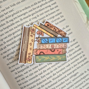 Op de afbeelding: Een sticker met een stapel geïllustreerde boeken met bloemen- en patroonontwerpen. De boeken zijn in verschillende kleuren, waaronder beige, oranje, groen en blauw. De sticker is geplaatst op een pagina van een boek.