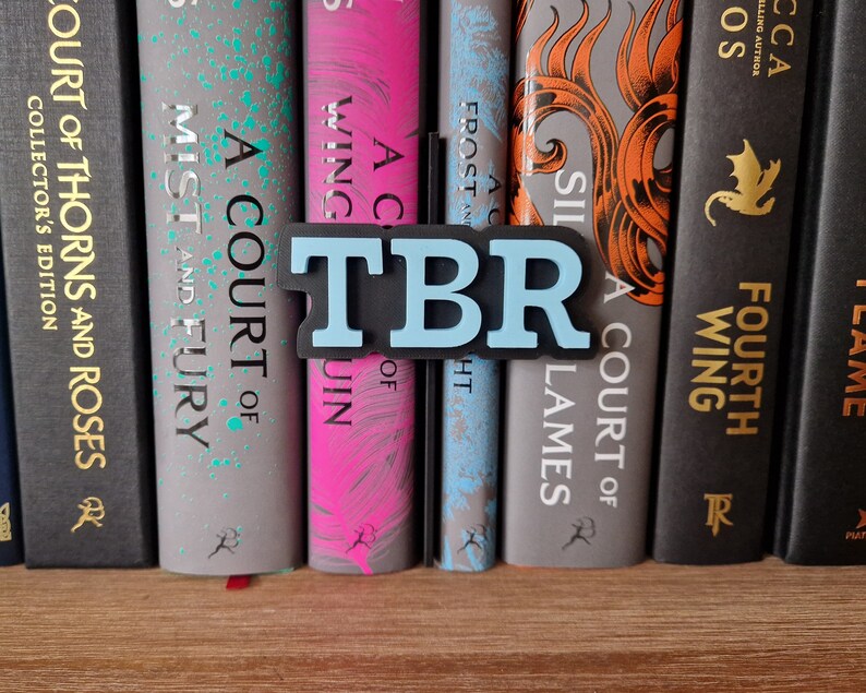May include: A bookshelf with a blue and black bookend that says "TBR" in large letters. The bookend is placed between two books with the titles "A Court of Wings and Ruin" and "A Court of Silver Flames".