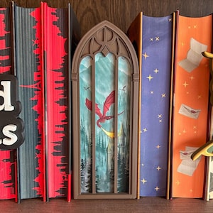 May include: A brown, arched, gothic-style book nook insert featuring a printed scene of a red dragon flying over a forest and a castle. The insert is positioned between several books with red, blue, and orange spines on a wooden shelf.