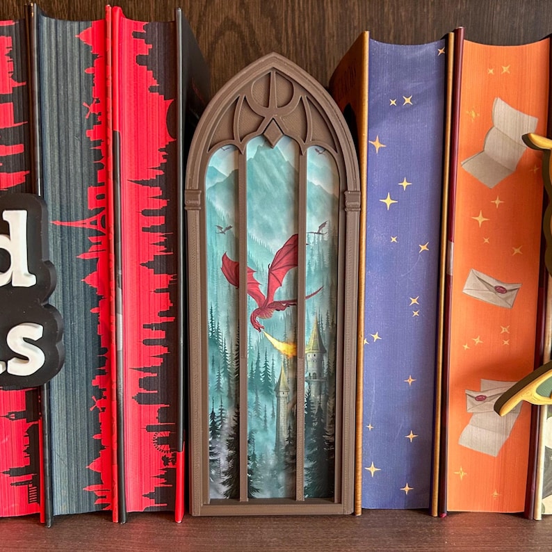 May include: A brown, arched, gothic-style book nook insert featuring a printed scene of a red dragon flying over a forest and a castle. The insert is positioned between several books with red, blue, and orange spines on a wooden shelf.