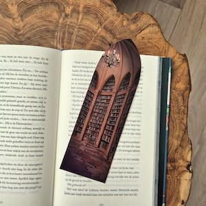 Op de afbeelding: Bladwijzer met een illustratie van een bibliotheekinterieur met gewelfde plafonds, boekenkasten en een kroonluchter. Het ontwerp heeft een donkerbruin en beige kleurenschema. De bladwijzer ligt op een open boek.