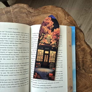 May include: A decorative bookmark with an illustration of a bookstore with autumn trees. The bookmark is placed on an open book. The bookstore has a sign that says "BOOKS". The bookmark has a curved top.