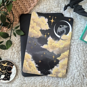 May include: A black notebook with a fantasy illustration. The design shows a dragon on a crescent moon, set against a backdrop of clouds and stars. The colours are black, gold, and cream.