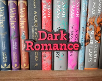 May include: A close-up of a bookshelf with a variety of books, including "A Court of Thorns and Roses" by Sarah J. Maas and "The Witcher" by Andrzej Sapkowski. A black and red sign with the words "Dark Romance" is placed in front of the books.