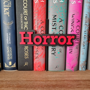 Op de afbeelding: Een rode en zwarte 3D-boekensteun met het woord "Horror". De boekensteun staat op een boekenplank met een verscheidenheid aan boeken, waaronder "A Court of Thorns and Roses" en "A Court of Mist and Fury".