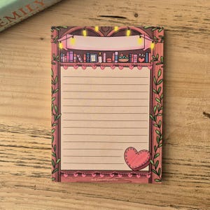 Könnte beinhalten: Ein Notizblock mit einem verspielten Design. Der Notizblock zeigt eine Bücherregal-Illustration mit Büchern, Lichterketten und einem Banner. Ein rosa Herz befindet sich in der unteren rechten Ecke, und die Ränder sind mit Blättern und Herzen verziert.