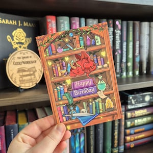 Op de afbeelding: Een kleurrijke verjaardagskaart met een boekenplankontwerp en een slapende rode draak. De kaart bevat de tekst "Happy Birthday" en is gevuld met fantasie-elementen. De kaart wordt in een hand gehouden, met een vage boekenplank op de achtergrond.