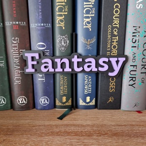 Op de afbeelding: Een paars en zwart bord met het woord "Fantasy" erop, geplaatst tussen een boekenkast met fantasyromans. De boeken bevatten titels als "The Witcher", "A Court of Thorns and Roses" en "A Court of Mist and Fury".