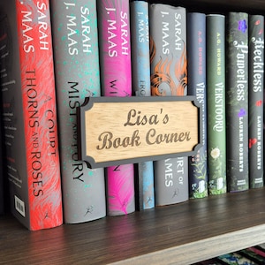Op de afbeelding: Een houten bord met de tekst "Lisa's Book Corner" staat voor een rij boeken op een plank. De boeken hebben kleurrijke covers, waaronder rood, roze en groen. Het bord heeft een donkergrijze rand.