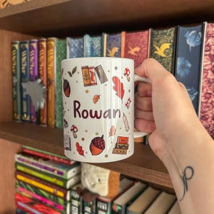Op de afbeelding: Witte keramische mok met de naam "Rowan" in bordeauxrood schrift, versierd met herfstthema-illustraties. De mok heeft kleurrijke bladeren, eikels en paddenstoelen. De mok staat op een houten plank met boeken.