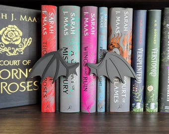 May include: A set of black dragon bookends with wings spread wide, holding up a row of fantasy novels with colorful covers. The books are titled "A Court of Thorns and Roses", "A Court of Mist and Fury", "A Court of Wings and Ruin", "A Court of Frost and Starlight", "A Court of Flames", "Versplinterd", and "Verstoord".