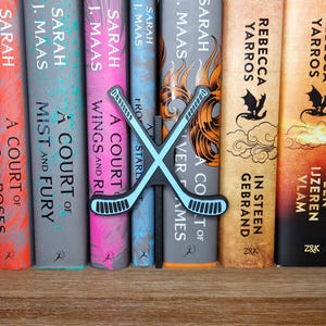 Puede incluir: Una estantería con una variedad de novelas de fantasía, incluyendo "A Court of Thorns and Roses" de Sarah J. Maas y "A Court of Mist and Fury" de Sarah J. Maas. Un marcapáginas de hockey azul y blanco está colocado entre los libros.