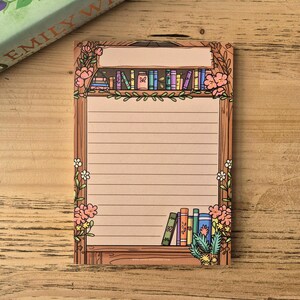 Peut inclure: Un bloc-notes ligné avec un motif de bibliothèque. Le bloc-notes présente un cadre en bois avec une étagère de livres colorés en haut et en bas. Des fleurs roses et des feuilles vertes décorent le cadre. Le bloc-notes est sur une surface en bois.