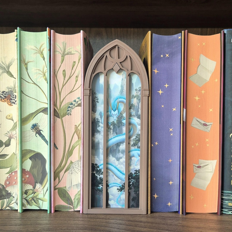 May include: A book nook insert featuring a light brown, arched gothic window frame. Inside the frame is a layered scene depicting a long, winding blue dragon flying through a mountainous landscape with clouds and dark green trees.