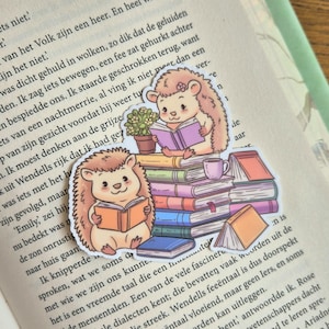 Op de afbeelding: Twee schattige egelstickers die boeken lezen, een op een stapel kleurrijke boeken met een kleine potplant en een theekopje. De stickers staan op een pagina van een boek met tekst.