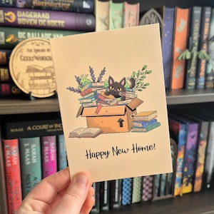 Op de afbeelding: Beige kaart met een cartoon illustratie van een zwarte kat in een doos vol boeken. De tekst "Happy New Home!" staat eronder. Op de achtergrond een boekenplank vol boeken.