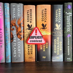 Op de afbeelding: Een boekenkast met een selectie fantasy-romans, waaronder "A Court of Thorns and Roses" van Sarah J. Maas, "Frost and Starlight" van Sarah J. Maas, "A Court of Silver Flames" van Sarah J. Maas, "A Court of Wings and Ruin" van Sarah J. Maas, "Steen" van Rebecca Yarros, "Een Eeren Lam" van Rebecca Yarros, "Door Duisternis Gesmeed" van Rebecca Yarros, "Powerless" van Lauren Roberts en "Reckless" van Lauren Roberts. Een rood en wit waarschuwingsbord met de tekst "Expliciete inhoud" is op de boekenkast geplaatst.