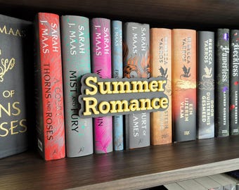 May include: A bookshelf displays a collection of books with colorful spines. A 3D sign that reads "Summer Romance" is placed in front of the books. The books are by authors like Sarah J. Maas and Rebecca Yarros.
