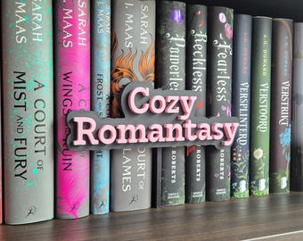 May include: A bookshelf with a collection of books, featuring titles like "A Court of Mist and Fury" and "Wings and Ruin." A pink and gray "Cozy Romantasy" bookend sits in front of the books. The books have colorful spines.