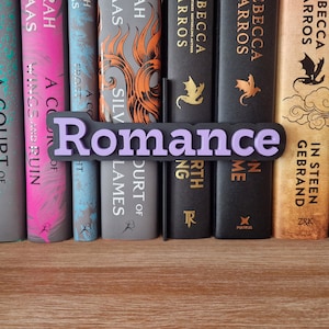 Puede incluir: Una estantería con una variedad de libros, incluyendo novelas de fantasía y romance. Los libros están dispuestos en una fila, con un letrero morado que dice "Romance" delante de ellos.