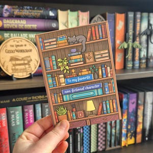 Op de afbeelding: Kleurrijke illustratie van een boekenplank met boeken, een kat en planten. De tekst op de kaart luidt "To my favorite non-fictional character". De kaart wordt voor een boekenplank vol boeken gehouden.