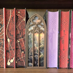 May include: A brown, arched, gothic-style book nook insert featuring a detailed illustration of a griffin perched on a rocky cliff overlooking a winding river valley, nestled between several hardcover books with red, black, and purple spines.