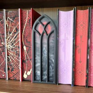Peut inclure: Un cadre noir de style gothique en forme d'arche contenant une illustration en niveaux de gris de loups hurlant à la lune rouge, placé entre plusieurs livres aux dos décoratifs avec des motifs rouges, noirs et violets, incluant des toiles d'araignée.