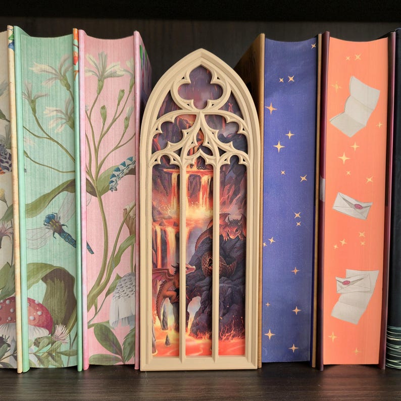 May include: A beige, arched, gothic-style book nook insert featuring a detailed scene of dragons in a volcanic landscape with flowing lava, positioned between two books with floral spines and two books with starry and envelope patterns.