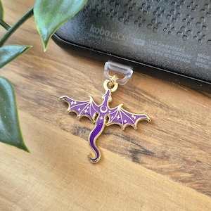 Puede incluir: Un dije de dragón morado con ribetes dorados y detalles de estrellas. El dragón tiene alas extendidas y una cola curvada. El dije está sujeto a un lazo de plástico transparente. El fondo incluye una superficie de madera y un dispositivo electrónico negro.