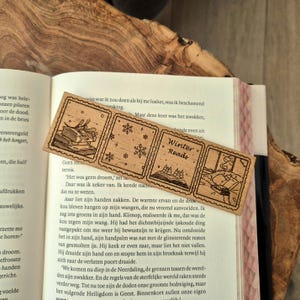 Op de afbeelding: Houten bladwijzer met vier rechthoekige panelen. De panelen bevatten illustraties van boeken, sneeuwvlokken, een winterlandschap en een lezende persoon. De tekst "Winter Reads" is ook aanwezig.