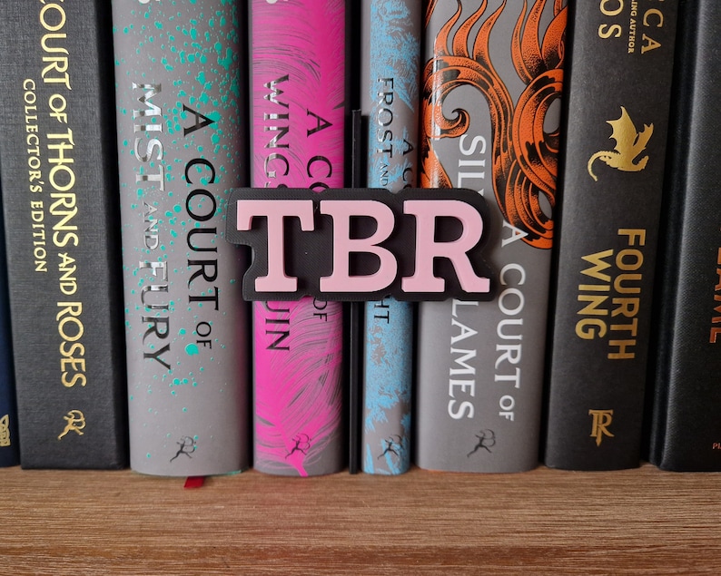 May include: A close-up of a bookshelf with a pink and black "TBR" book marker. The bookshelf is filled with fantasy novels with titles like "A Court of Thorns and Roses" and "A Court of Mist and Fury".