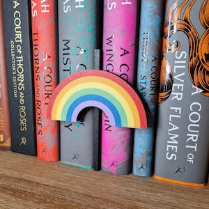 Op de afbeelding: Een regenboogkleurige boekenlegger in de vorm van een regenboog. De boekenlegger staat tussen een rij boeken met titels als "A Court of Thorns and Roses" en "A Court of Silver Flames".