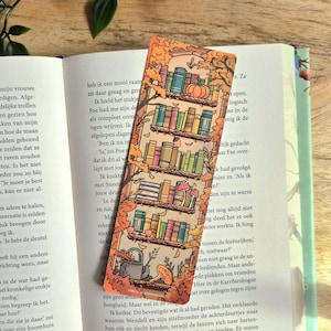 Op de afbeelding: Een rechthoekige boekenlegger met een verticale illustratie van een boekenplank met vijf niveaus in een herfstbos. De planken bevatten kleurrijke boeken, een pompoen, paddenstoelen en een gieter, omlijst door oranje en bruin herfstblad.