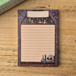 May include: A lined notepad with a fantasy theme. The design features a bookshelf with books, bottles, and a skull. The notepad has a purple border with a blank label at the top. The bottom of the notepad has two bottles and a skull.