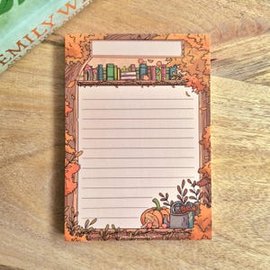 Op de afbeelding: Een rechthoekig notitieblok met een gelinieerd schrijfgedeelte, omlijst door een herfstthema met oranje bladeren, een plank met kleurrijke boeken, een pompoen en een gieter. Bovenaan de pagina bevindt zich een leeg rechthoekig tekstvak.